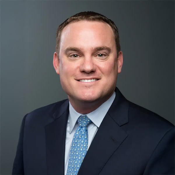 Headshot of Elliott Throne, a man with short brown hair and a friendly smile, wearing a dark suit jacket, light blue shirt, and patterned blue tie, against a plain gray background.