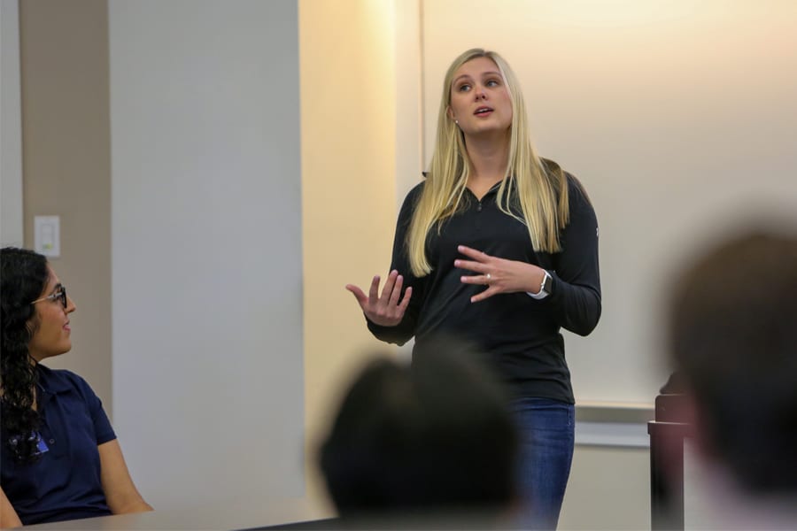 A young woman with long blonde hair speaks animatedly in front of a small group in a classroom setting, while a seated woman watches attentively.