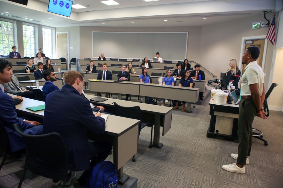 A student stands at the front of a classroom speaking to an attentive group of students seated in tiered rows.