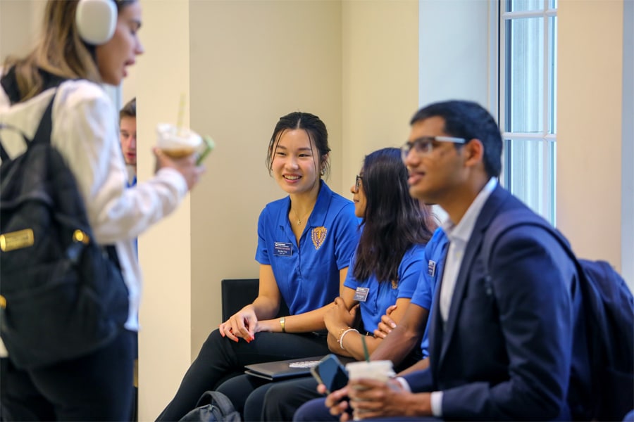 Three students in blue Warrington polo shirts sit chatting and smiling near a window while another student wearing headphones and holding a drink walks past.