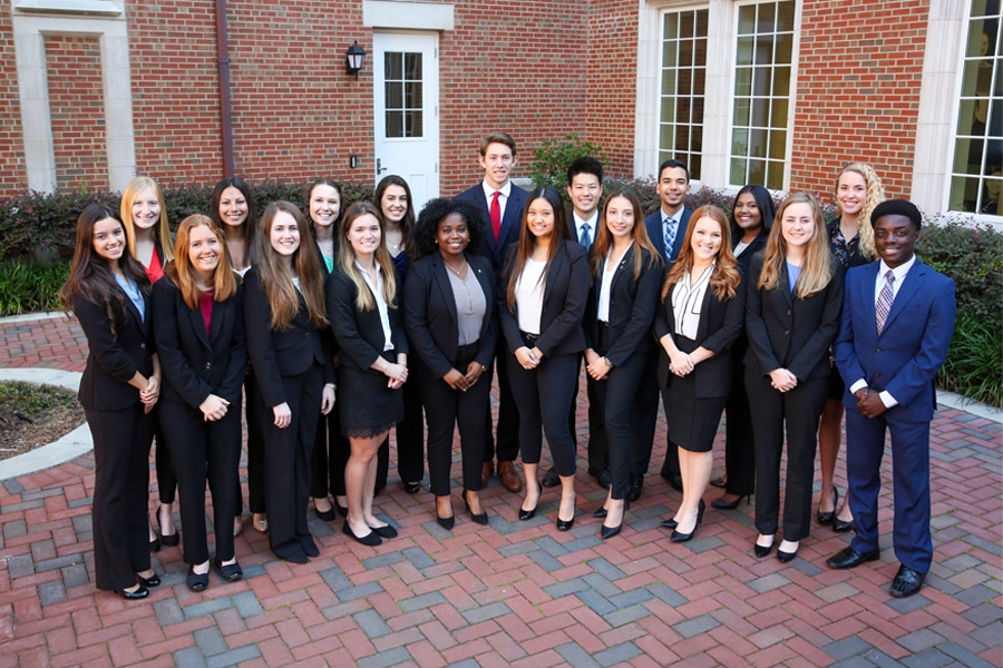 A diverse group of young professionals dressed in business attire pose together outside a brick building, smiling confidently toward the camera.