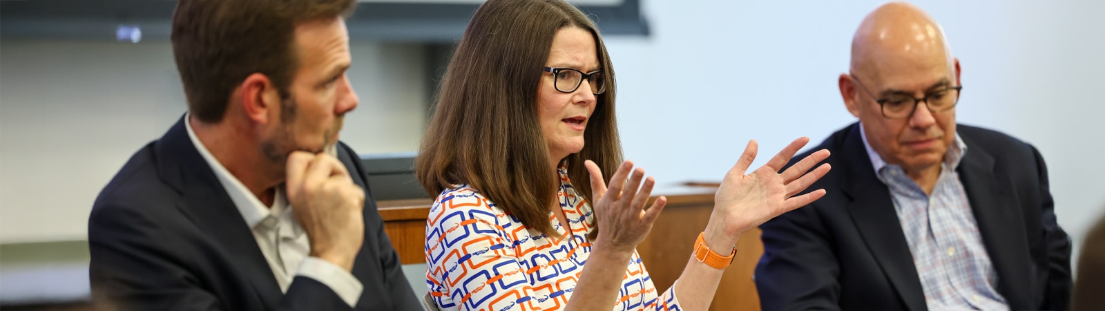A woman gestures while speaking between two men seated beside her during a professional finance speaker panel discussion.