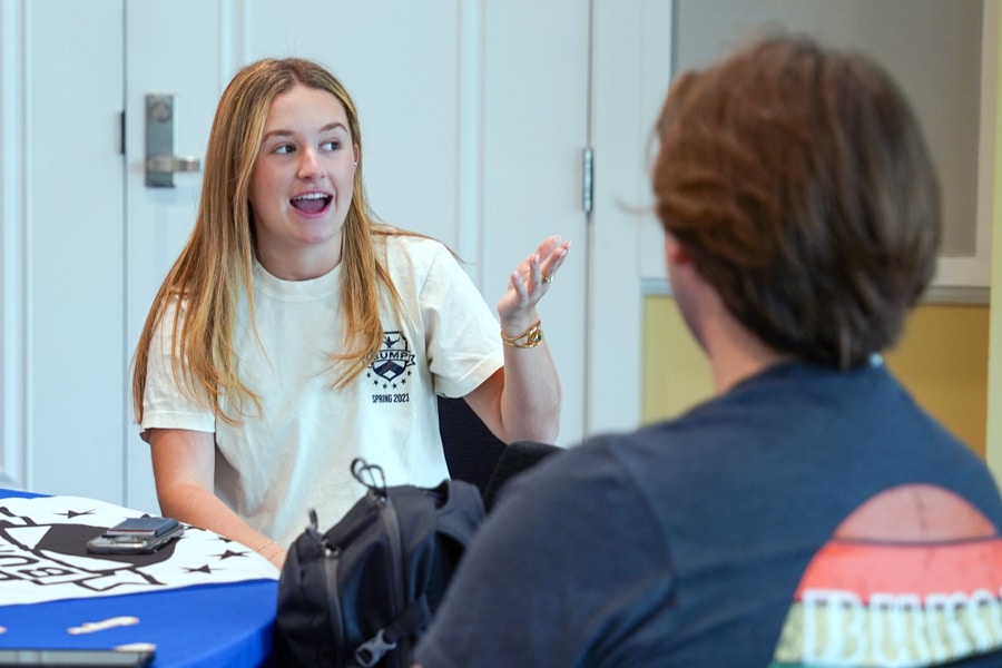 A female student wearing a white BUMP t-shirt speaks animatedly to a male student seated across from her in a casual setting with a table and backpacks nearby.