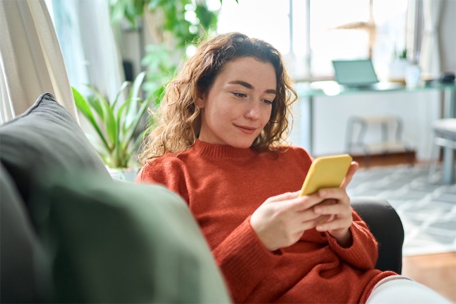 A student lounges on a couch at home, looking at her smartphone screen with a focused expression.