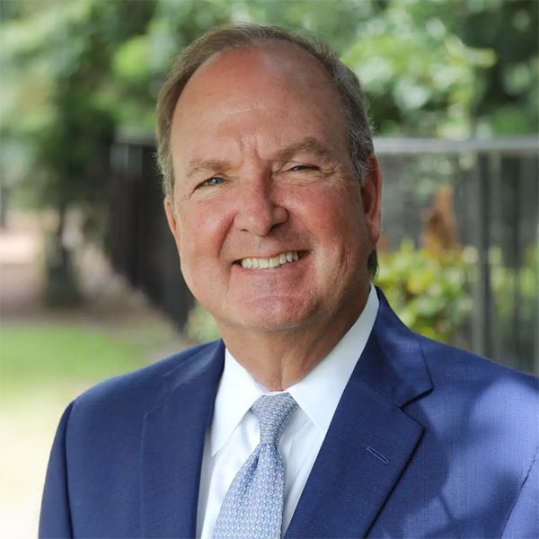 Headshot of Gilbert Pomar, a man with light brown hair and a warm smile, wearing a blue suit, white shirt, and light blue patterned tie, with a blurred outdoor background.