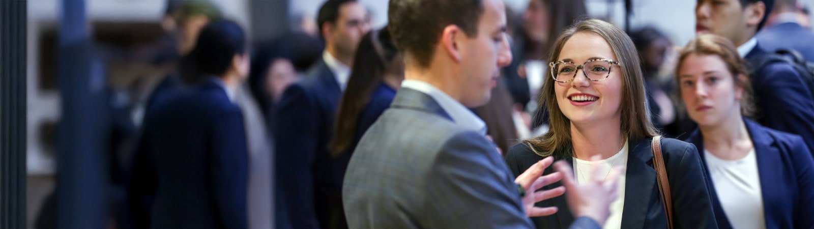 A young woman in business attire smiles and listens attentively during a conversation with a male professional in a bustling networking event or career fair setting.