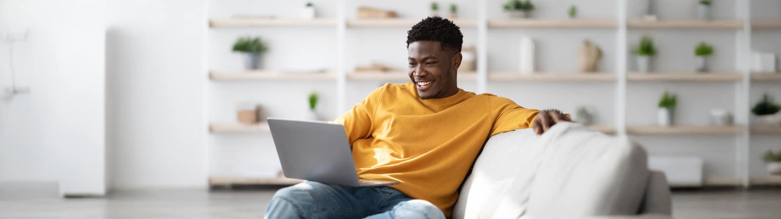 A young man in a mustard yellow sweater smiles while using a laptop, seated comfortably on a light gray couch in a bright, modern living room.