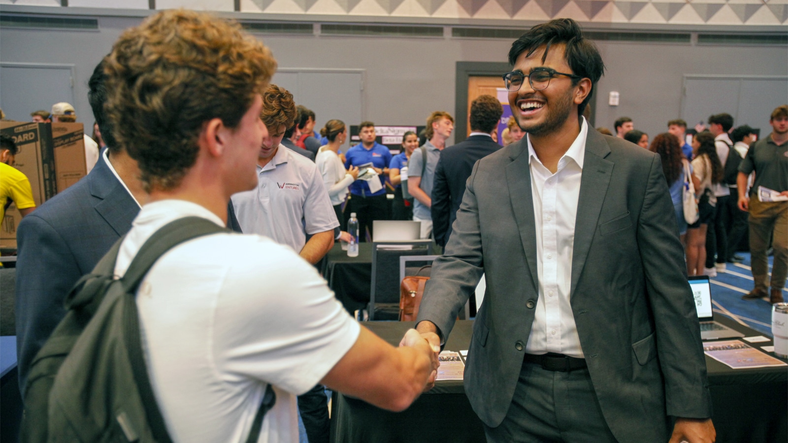 A student in business attire smiles and shakes hands with a peer at a busy on‑campus career fair table surrounded by other students.