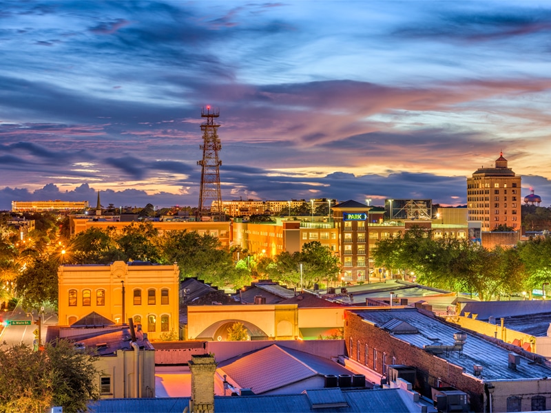 A vibrant view of downtown Gainesville at sunset, featuring a mix of modern and historic buildings, a tall radio tower, and trees lining the streets. The sky glows with shades of orange, blue, and purple.