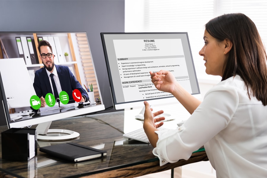 A woman reviews a resume on one computer monitor while video conferencing with a man in business attire on a second monitor.