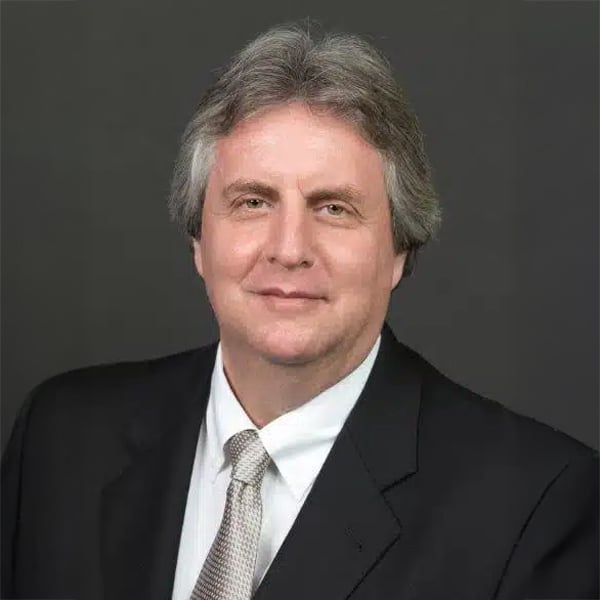Professional headshot of Howard Gutman, a middle-aged man with gray hair, wearing a black suit, white shirt, and light gray patterned tie, against a dark gray background.