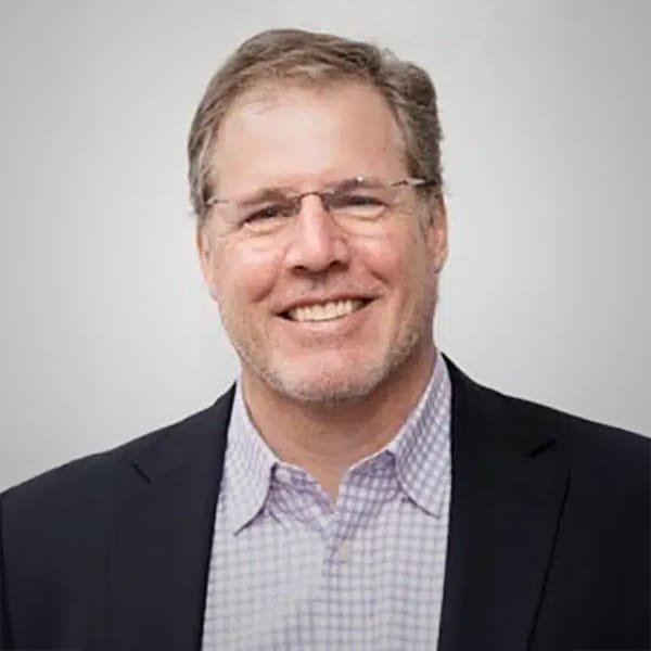 Portrait of Joe Brady, a middle-aged man with light brown hair and glasses, wearing a dark blazer over a light checkered shirt, smiling against a plain white background.