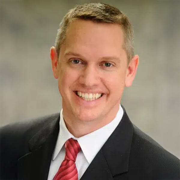 Headshot of John Crossman, a smiling man with short light brown hair, wearing a dark suit jacket, white shirt, and red tie, posed against a neutral background.