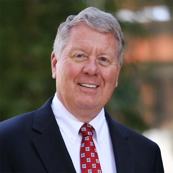 Portrait of Kelley A. Bergstrom, a man with gray hair, wearing a dark suit jacket, white dress shirt, and a red patterned tie, smiling warmly outdoors with trees softly blurred in the background.