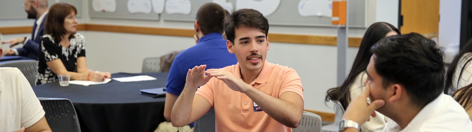 A student in a peach polo shirt leads a discussion with peers at a round table during a leadership workshop. He gestures as he speaks, while others listen attentively in a collaborative classroom setting.