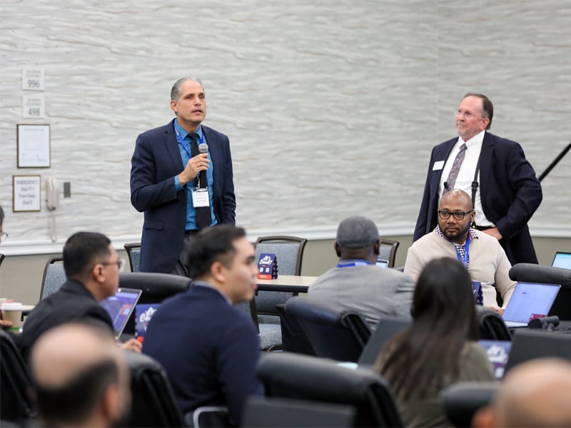 A professional stands and speaks with a microphone during a PURC training session, while another facilitator looks on and participants listen in a conference room setting.
