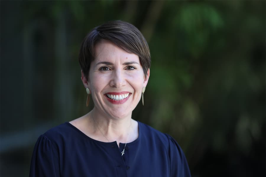 Headshot of Lisa Dilts, a smiling woman with short brown hair, wearing a navy blue top and gold earrings, standing outdoors with blurred greenery in the background.