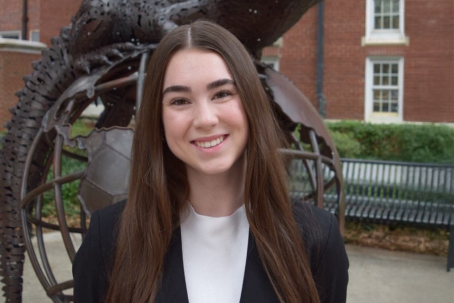 Mackenzie Nichols, VP of Internal Operations for Business Ethics Ambassadors, smiles in a professional outdoor headshot with long brown hair and a black blazer, standing in front of a globe sculpture.