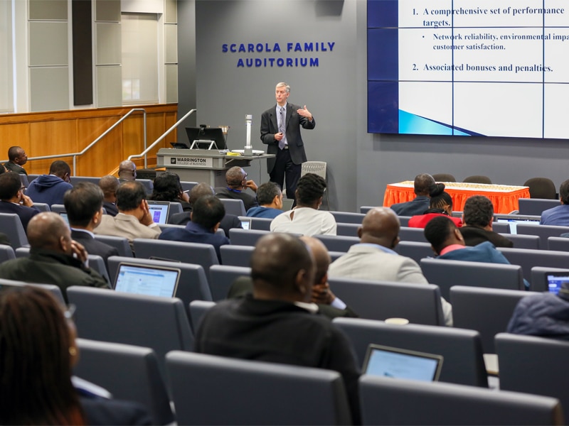 A speaker delivers a presentation on utility performance management to a group of professionals in the Scarola Family Auditorium, with attendees seated in rows and following along on laptops.
