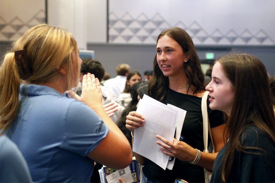 A young female student wearing a black top holds papers and listens attentively as a student in a blue polo shirt speaks energetically to her at a crowded event.