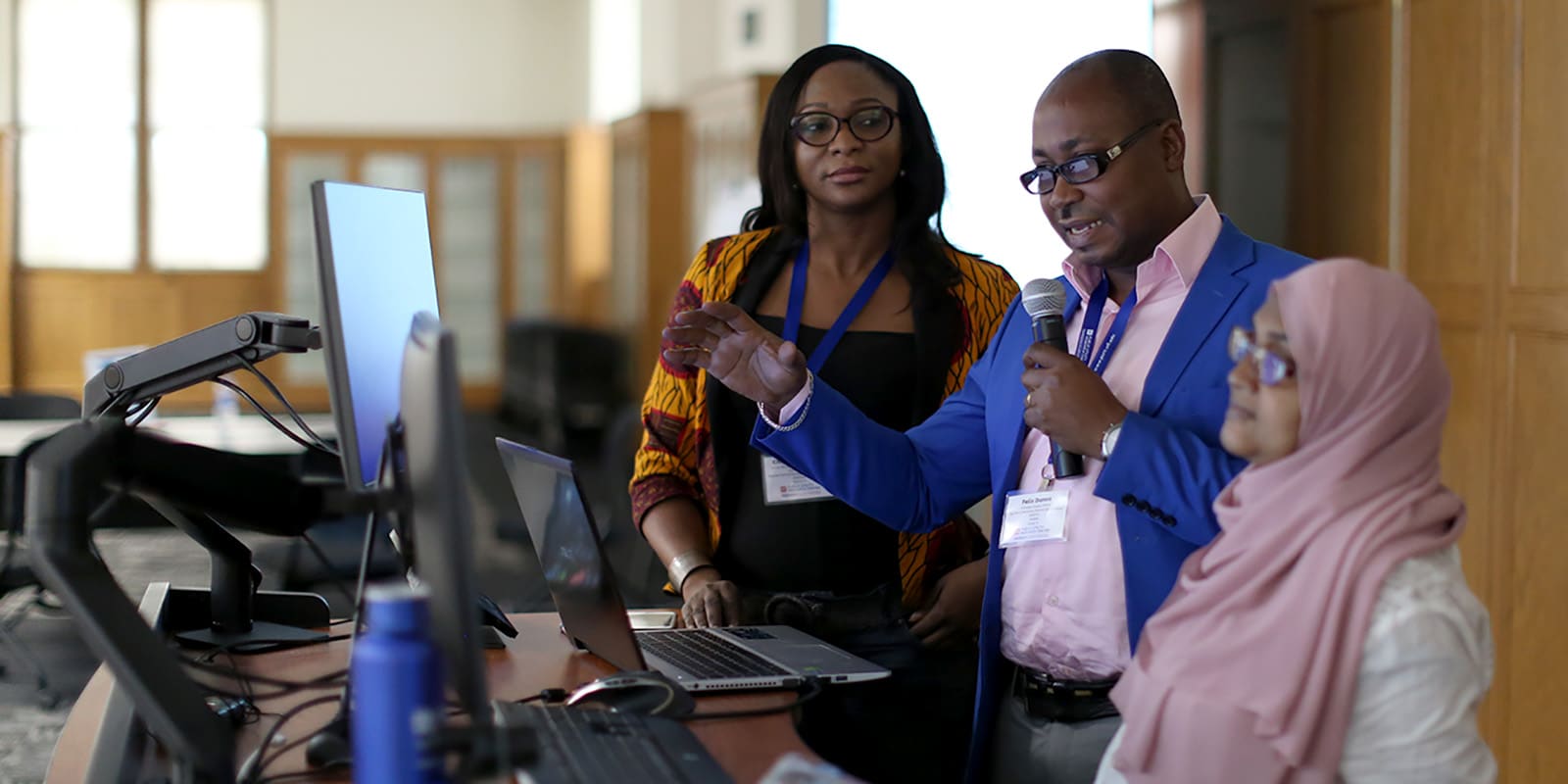 Group of people standing at a computer with the person in the middle holding a microphone and talking.