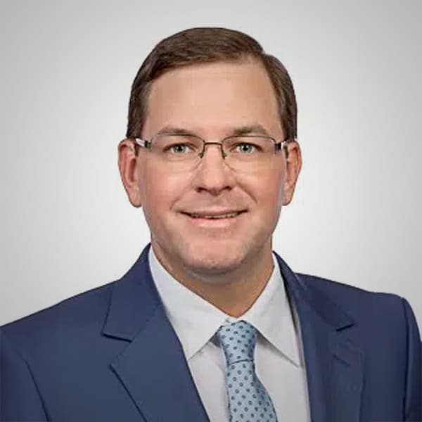 Portrait of Patrick Nutt, a man with short brown hair and glasses, wearing a blue suit, white shirt, and light blue patterned tie, smiling against a plain gray background.