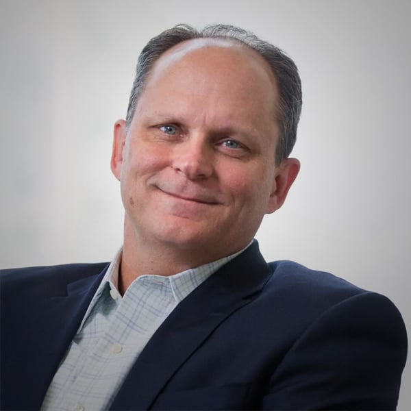 Portrait of Paul Ellis, a middle-aged man with short hair and blue eyes, wearing a light-colored checkered shirt and navy blazer, smiling gently against a white background.