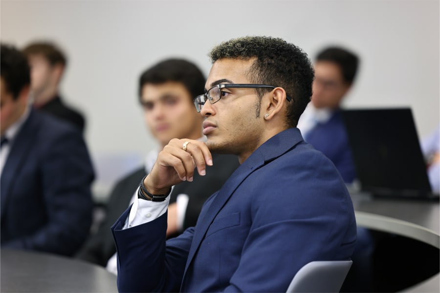 A young man in a navy blue suit and glasses listens thoughtfully with his hand resting near his chin in a classroom setting.