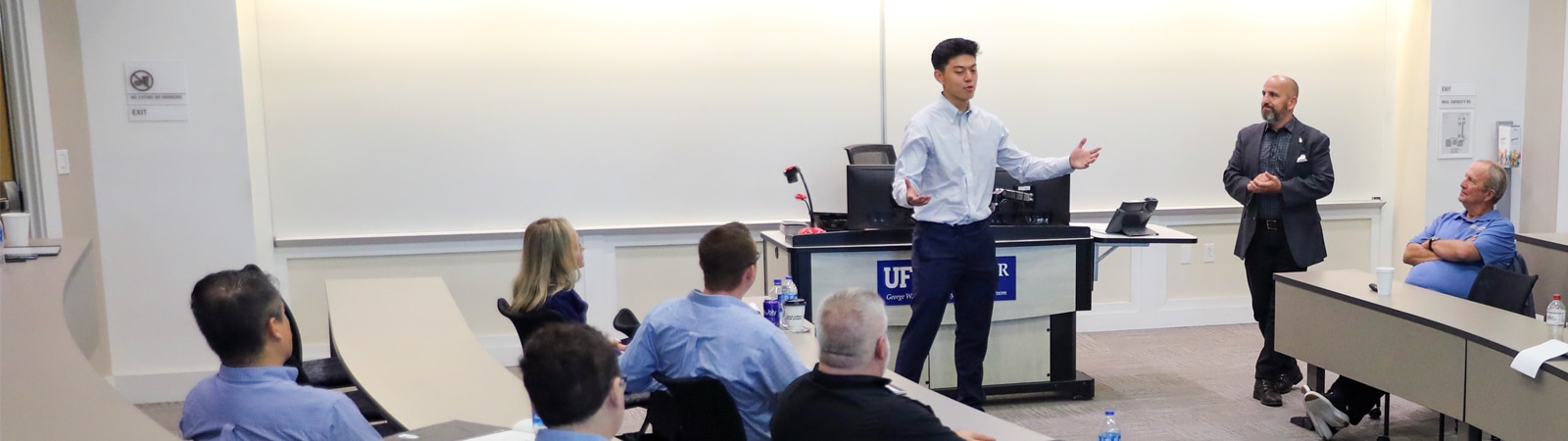A student stands at the front of a business classroom, presenting to an audience of faculty and peers, while a professor observes from the side near a UF Heavener School of Business podium.