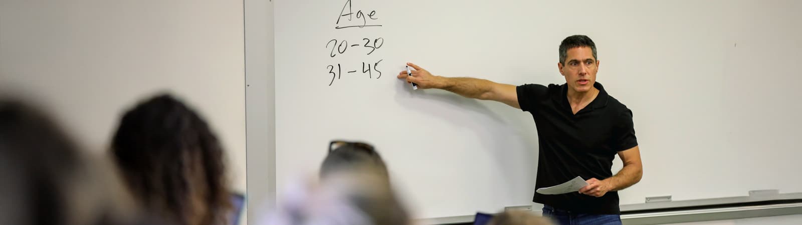 Aner Sela, City Furniture Professor in the Marketing Department at Warrington College of Business, stands at the front of a classroom, pointing to a whiteboard with age ranges written on it, as students listen attentively.