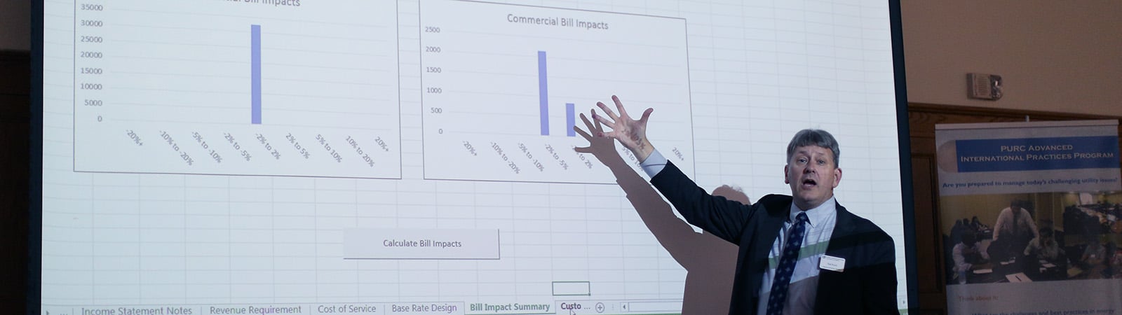 Older man standing and gesturing in front of a presentation with data.