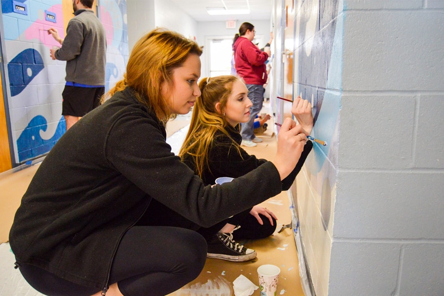 A group of young people, mostly female, kneeling and painting a colorful mural on a hallway wall, with additional students painting further down the corridor.