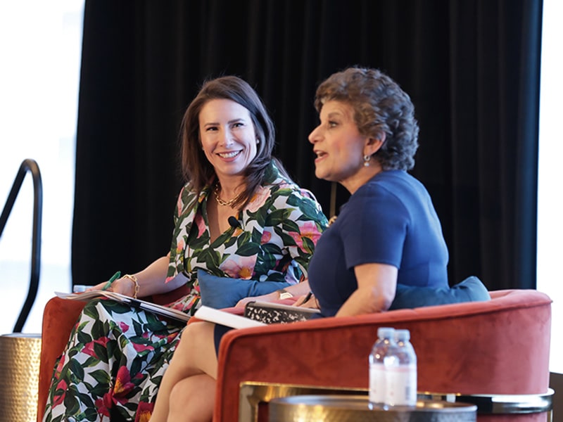 Two women seated on stage talking to each other and an audience at a conference.
