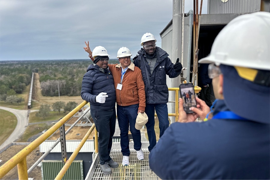 Three utility professionals wearing hard hats and safety gear pose for a photo atop a high platform at an industrial facility, while a fourth person takes their picture. A broad landscape and railroad track are visible in the background.
