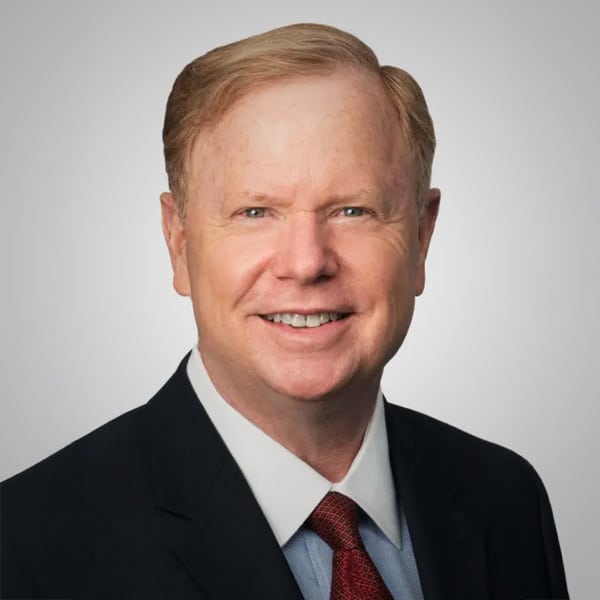 Portrait of Richard Bezold, a middle-aged man with light red hair, wearing a dark suit, light blue dress shirt, and a red patterned tie, smiling against a plain white background.