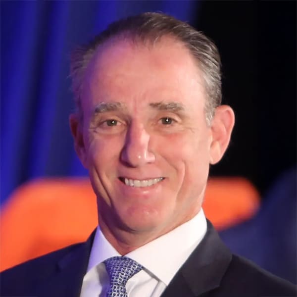 Headshot of Rick Scarola, a smiling man with short gray hair, wearing a dark suit jacket, white shirt, and patterned tie, against a blurred dark background with orange and blue hues.