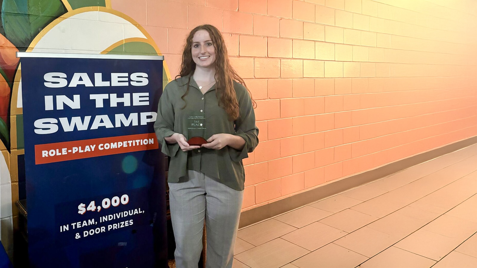 A student stands smiling and holding a 1st place award in front of a banner for the "Sales in the Swamp" role-play competition, with prize information and a colorful mural in the background.