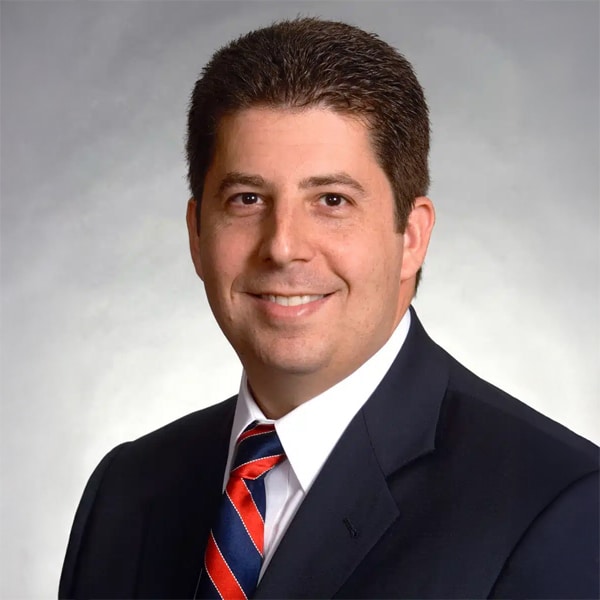 Professional headshot of Seth Wise, a man with short dark hair, wearing a dark suit jacket, white shirt, and a striped red and blue tie, smiling against a neutral gray background.