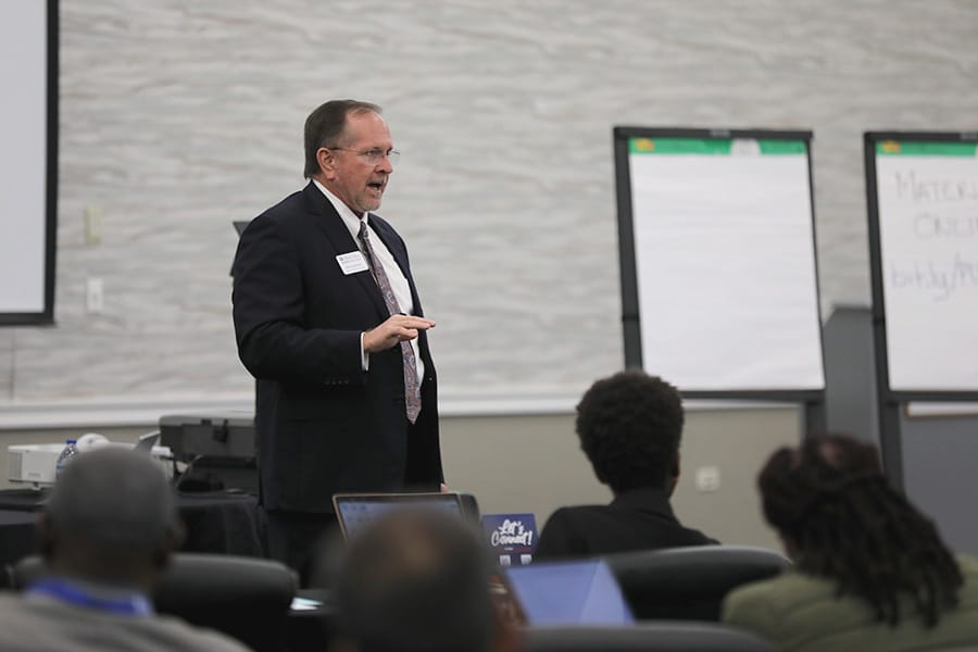 Older man standing and speaking at a conference with people sitting in the audience in front of him.