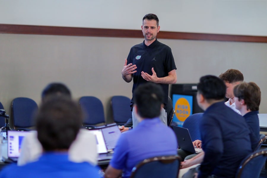 A speaker in a black UF Gators shirt stands addressing a group of students or professionals seated around laptops, engaging them in discussion at a collaborative workshop.