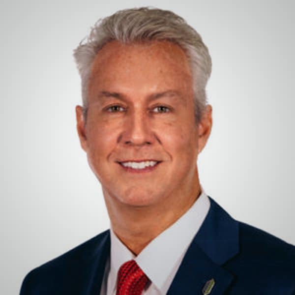Professional headshot of Stephen Hagenbuckle, a middle-aged man with light gray hair, wearing a dark blue suit, white shirt, and red tie, smiling against a light gray background.