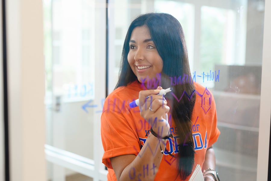 A young, smiling woman in a University of Florida t-shirt writing on a clear dry-erase board.