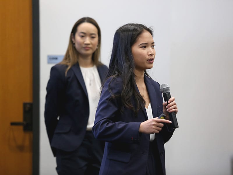Young woman in business attire presenting with another woman, her team member, in the background behind her.