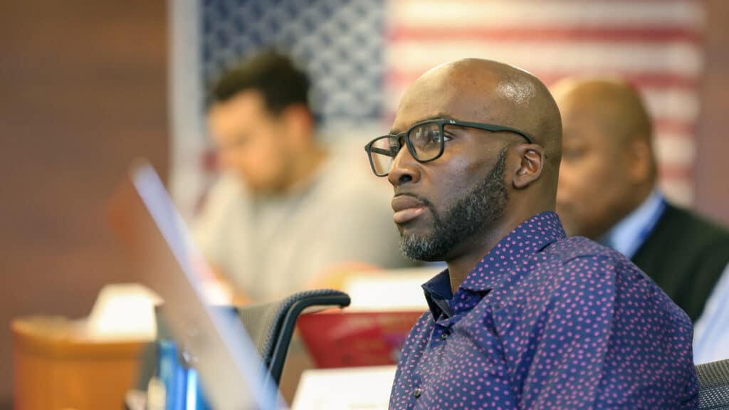 A focused, thoughtful man in a classroom listens intently, with an American flag blurred in the background. He wears glasses and a navy shirt with small red polka dots.