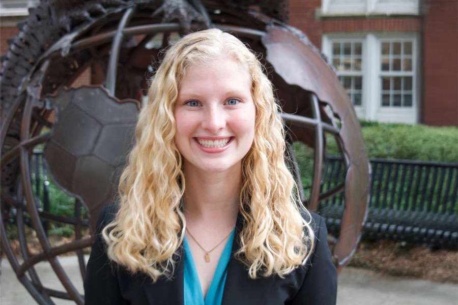 Taylor Aks, VP of Professional Development for Business Ethics Ambassadors, smiles in an outdoor portrait with long curly blonde hair, wearing a teal blouse and black blazer.