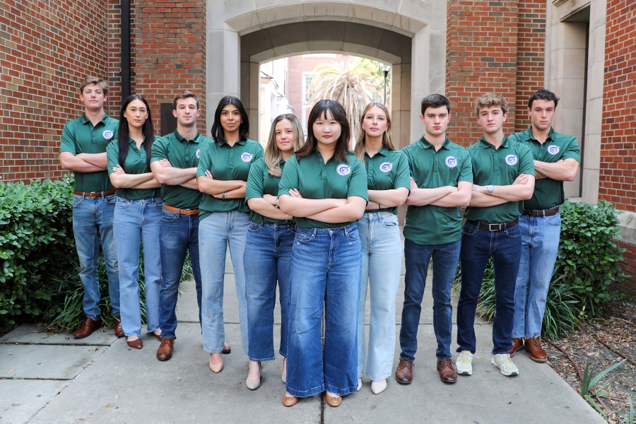 A group of 10 diverse undergraduate students stand in two rows outdoors between brick buildings, all wearing matching green polo shirts and jeans, arms crossed confidently.