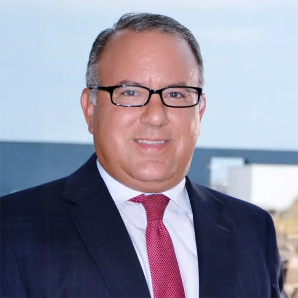 Headshot of Timothy Peterson, a man with short dark hair and glasses, wearing a dark suit, white shirt, and red patterned tie, smiling with a blurred outdoor background.