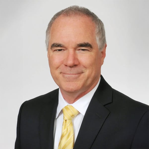 Professional headshot of Todd Jones, a middle-aged man with short gray hair, wearing a black suit, white shirt, and yellow striped tie. He smiles gently against a plain light background.