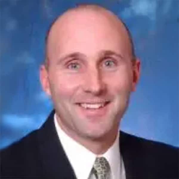 Headshot of Todd Powell, a smiling man wearing a dark suit, white shirt, and patterned tie, set against a soft blue background.