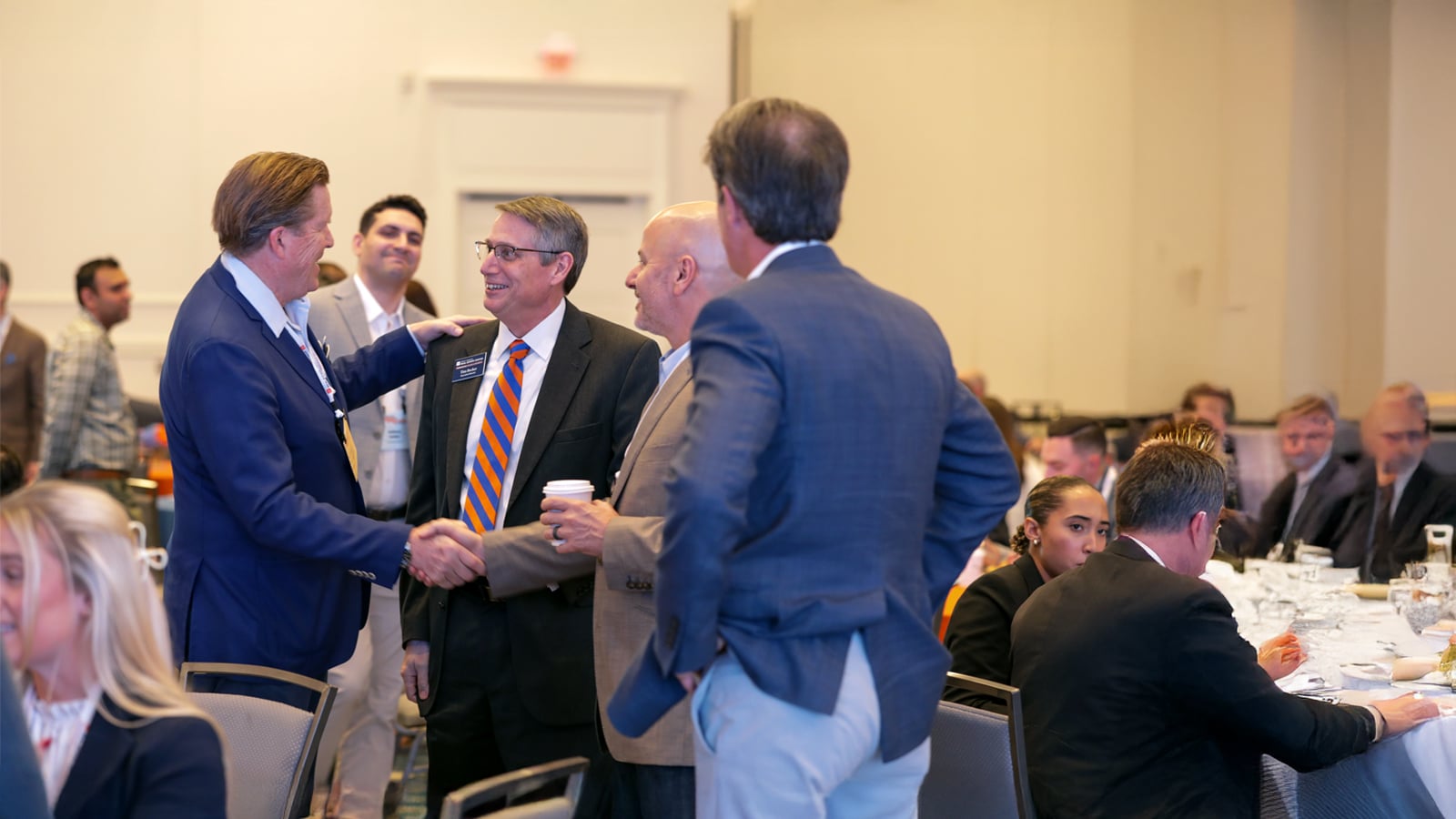 A group of well-dressed professionals greet and shake hands at a formal event, with tables and attendees in the background.